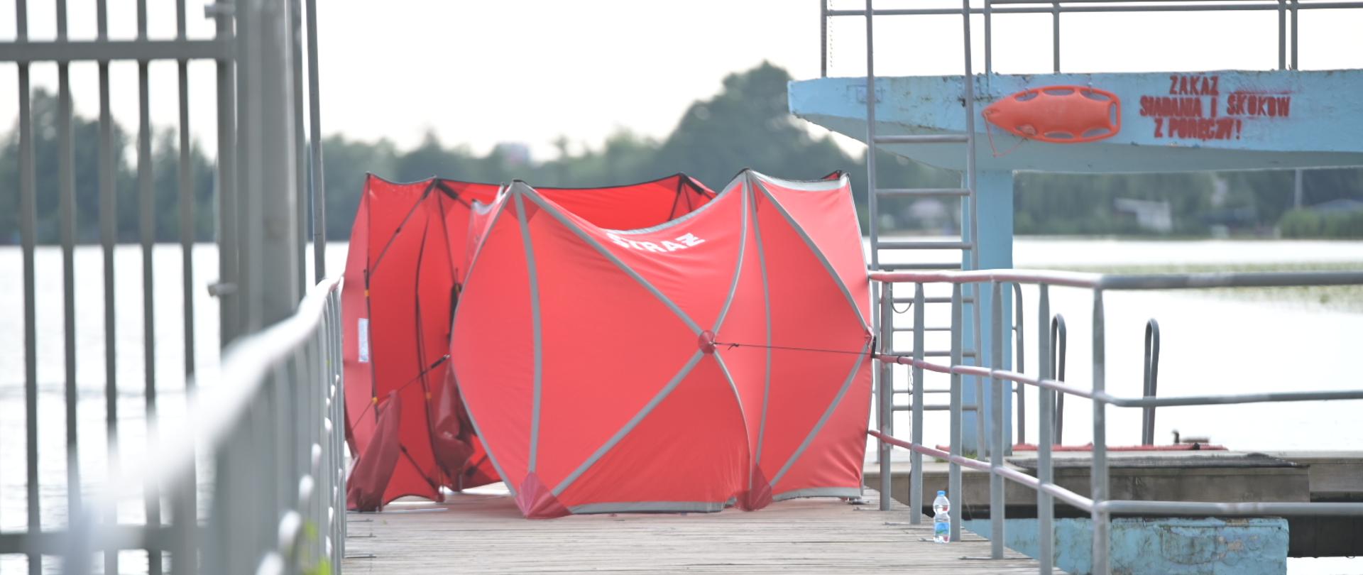 Na zdęciu widoczny czerwony parawan z napisem straż stojący na drewnianym pomoście przy brzegu jeziora.