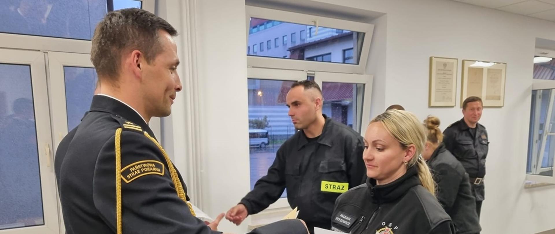 Na zdjęciu w sali konferencyjnej Komendy Powiatowej Państwowej Straży Pożarnej widoczny komendant powiatowy PSP w Gorlicach ubrany w ubranie galowe koloru granatowego ze sznurem wręczający zaświadczenia ukończenia szkolenia podstawowego strażaków ratowników OSP druhnie ubranej w ubranie koszarowe w czarnym polarze.