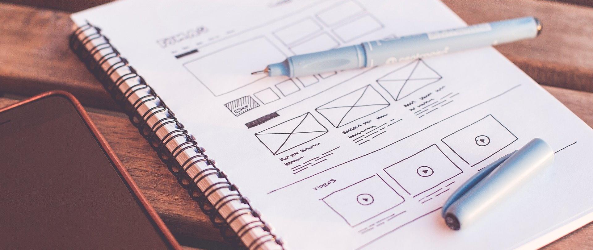 Na drewnianym stole leży kołozeszyt z zapełnioną pismem i ikonami pierwszą stroną. Na nim leży cienkopis w kolorze czarnym ze zdjętą skuwką. Z prawej strony kołozeszytu wyłączony telefon komórkowy.
