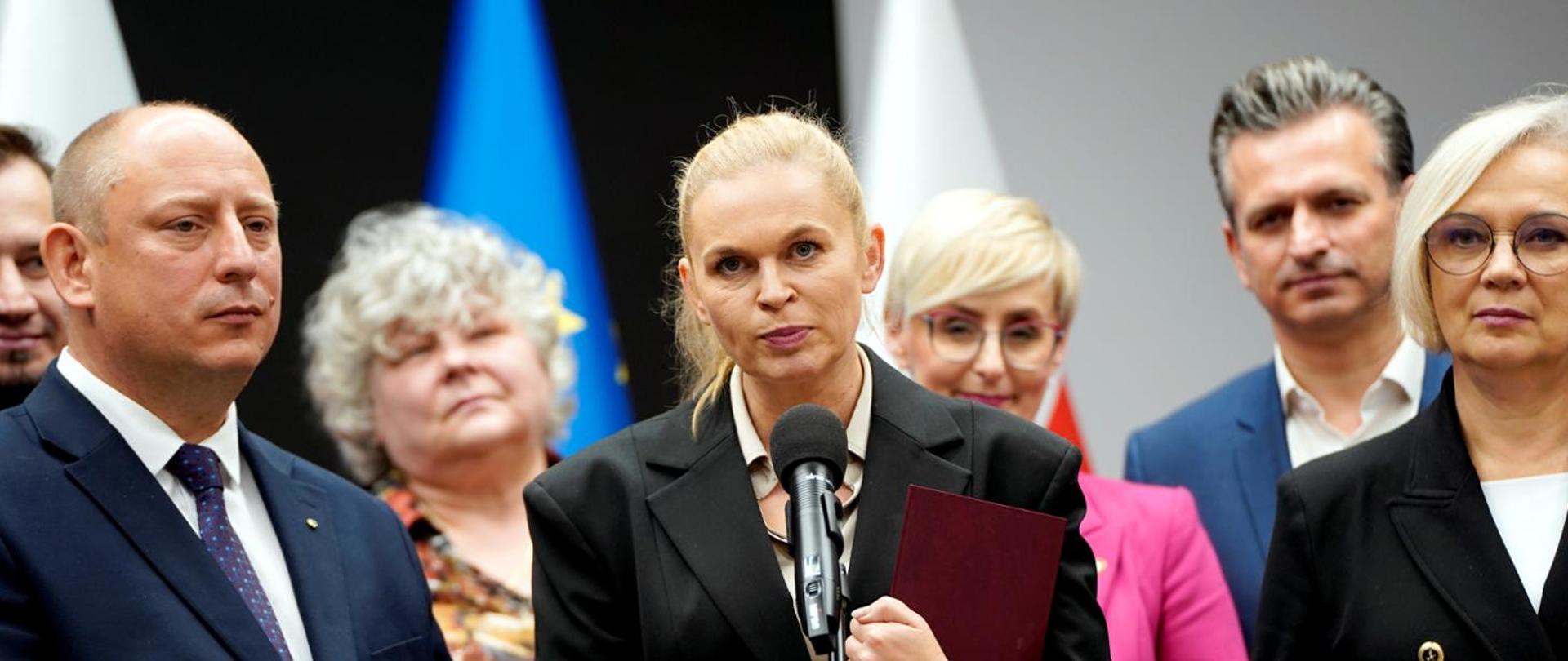 Minister Nowacka stoi przed mikrofonem na stojaku, w ręku trzyma fioletową teczkę, dookoła niej kilka osób.