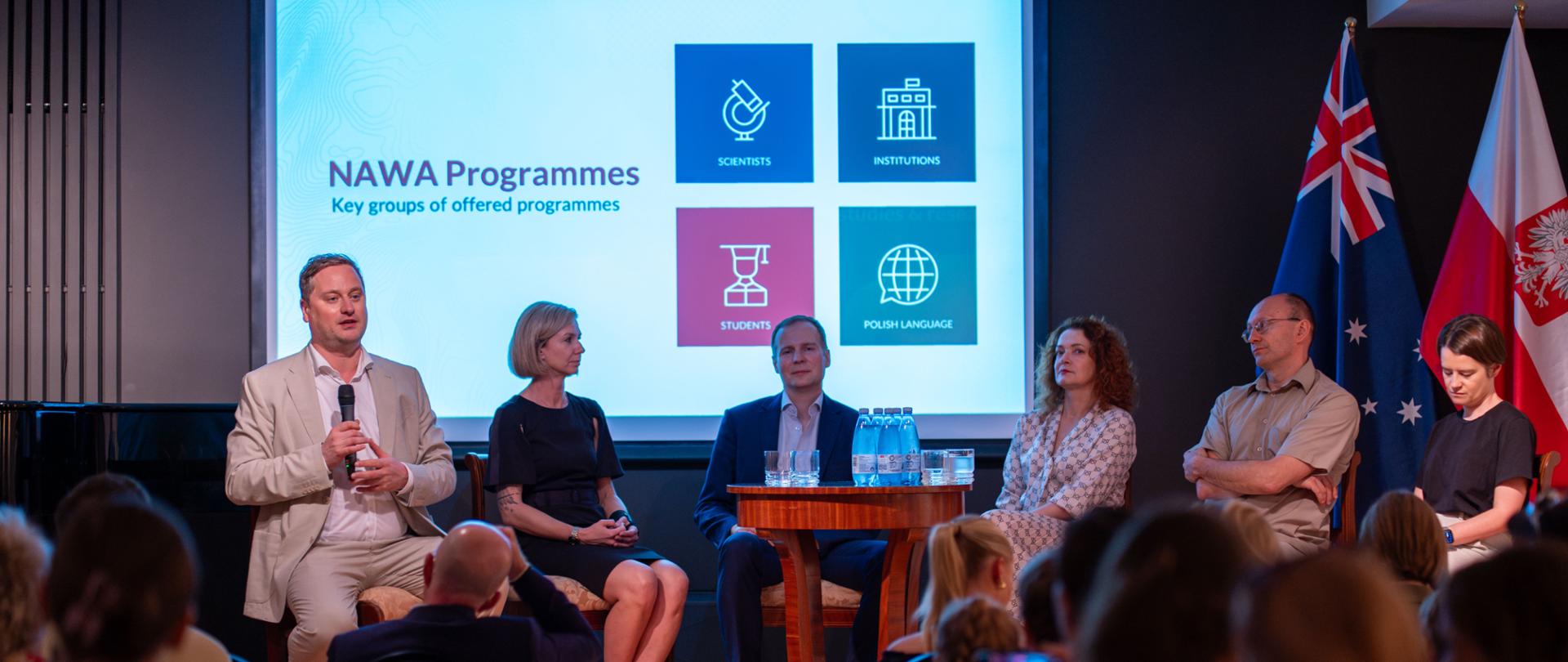 The photo shows six people sitting on a stage, one of them holding a microphone, with the Polish, Australian, and European Union flags in the background.
