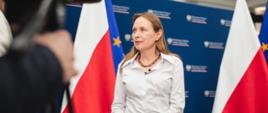 Minister Katarzyna Pełczyńska- Nałęcz stoi skierowana w lewą stronę przed dziennikarzem z kamerą.