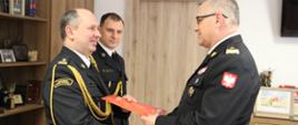 Trzech oficerów pożarnictwa w mundurach galowych. Generał wręcza akt powołania i zwraca się w kierunku strażaka. Drugi słucha. W tle szafa ze statuetkami.