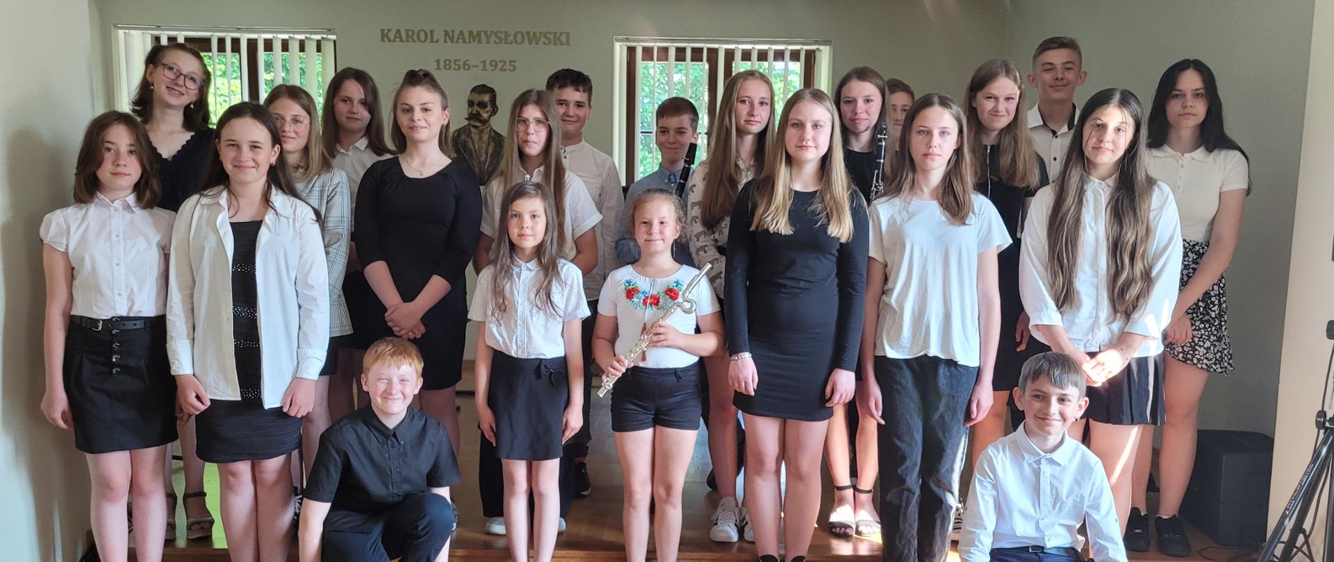 Sala koncertowa, przodem do publiczności ustawione dzieci -22osoby. Dzieci ubrane w stroje galowe. Dziewczynka stojąca pośrodku trzyma w dłoniach instrument -flet. Dwóch chłopców - jeden po lewej i jeden po prawej stronie kuca. W dali widać dwa okna oraz napis na ścianie Karol Namysłowski 1856-1925