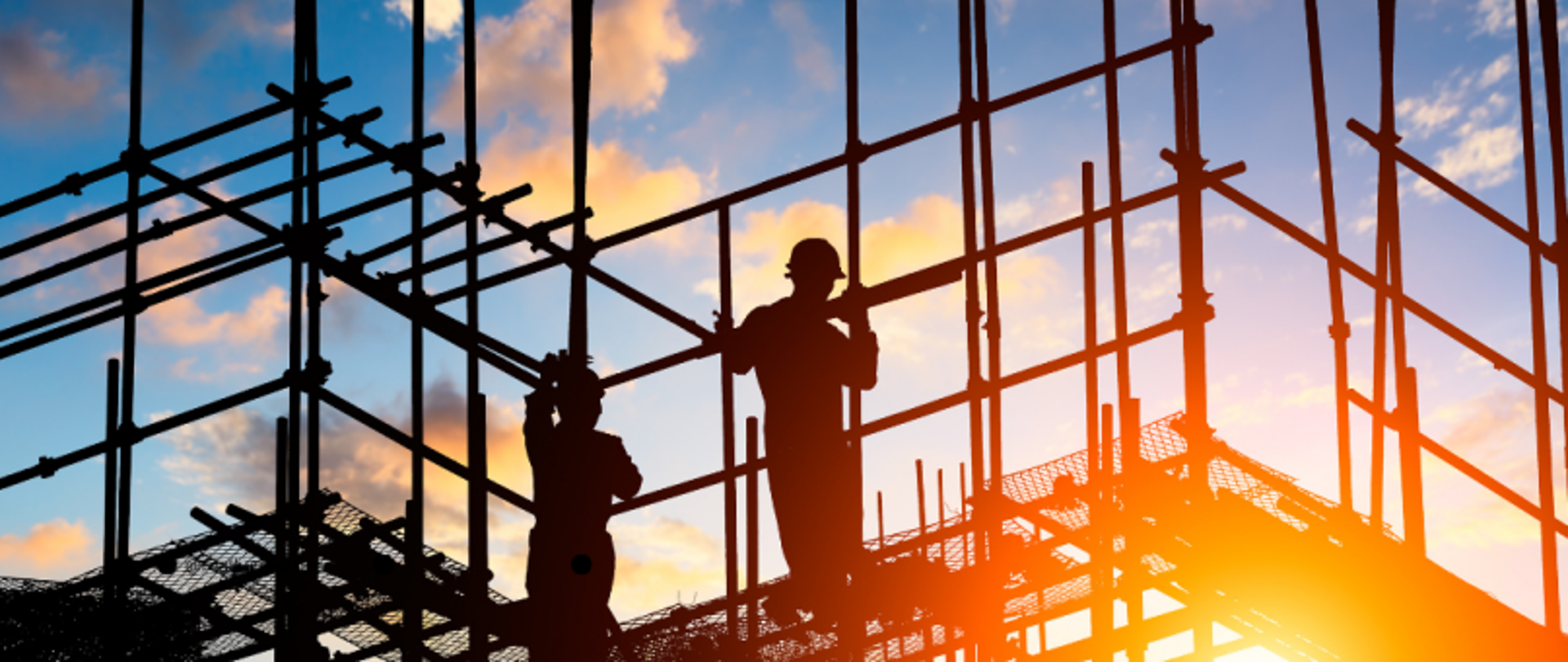 Dwóch pracowników budowlanych na rusztowaniu, sylwetki na tle nieba o wschodzie lub zachodzie słońca.

