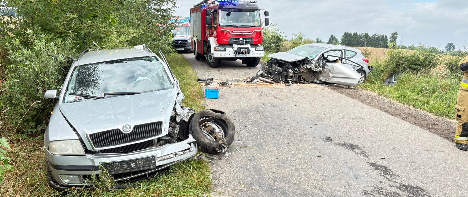 Dwa rozbite auta stojące na poboczu. W tle wóz PSP i ambulans ZRM.
