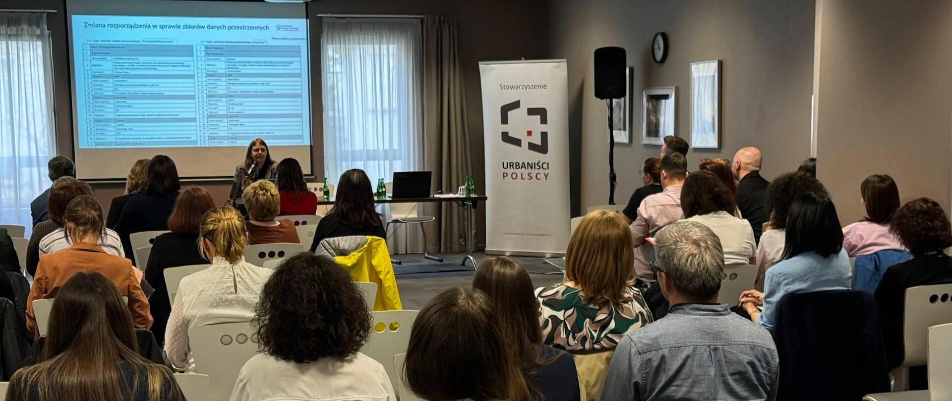 Po prawej stronie znajduje się baner reklamowy Stowarzyszenia Urbanistów Polskich oraz wysoki głośnik. W części centralnej za stołem prezydialnym siedzi Zastępca Dyrektora Departamentu Planowania Przestrzennego, Anna Michalik, która jest Prelegentem. na statywie umieszczony jest mikrofon, do którego przemawia prelegentka. W tle widoczny jest projektor, na którym wyświetlane są slajdy prezentacji. W dolnej części zdjęcia widoczni są uczestnicy seminarium, którzy siedzą na białych krzesłach.
