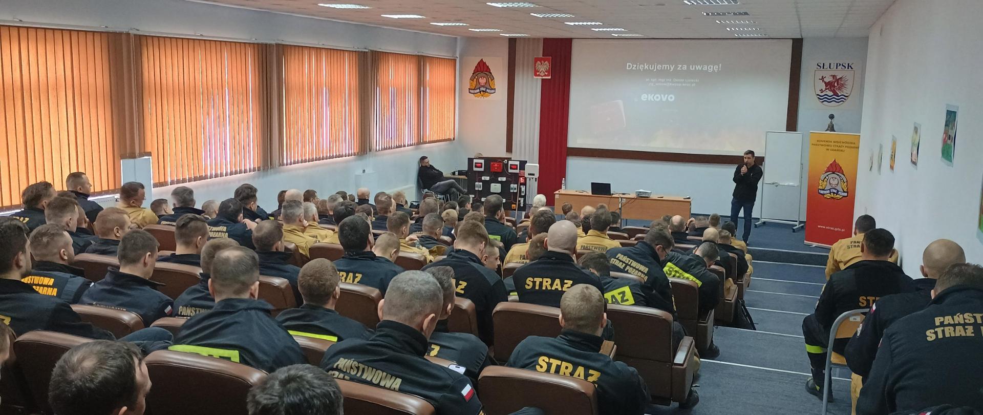 Strażacy siedzą na auli w krzesłach przed nimi stoi mężczyzna z mikrofonem obok stoi baner Komendy Wojewódzkiej PSP w Gdańsku na ekranie wyświetlana jest prezentacja. 