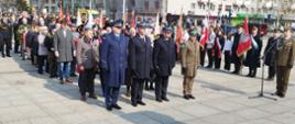 Delegacja złożona z czterech funkcjonariuszy służb mundurowych od lewej Policji, Straży Pożarnej oraz Wojska Polskiego stojąca na placu podczas uroczystości, za nimi stojący cywilni uczestnicy uroczystości