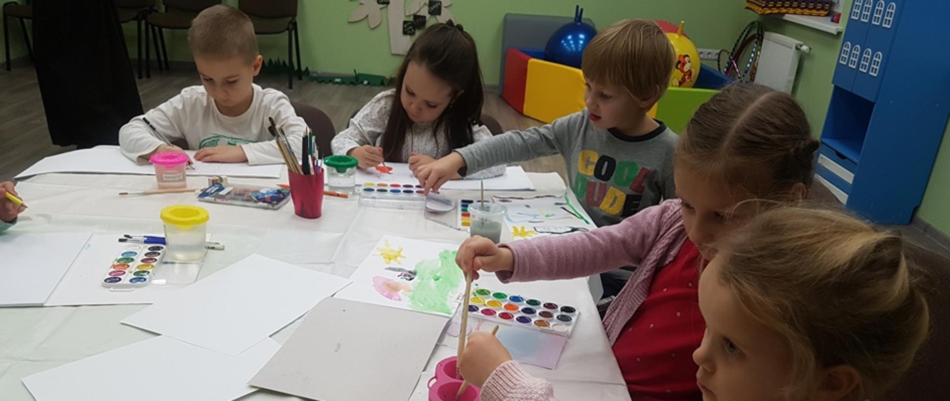 Dzieci siedzące przy stole malują kolorowymi farbkami 