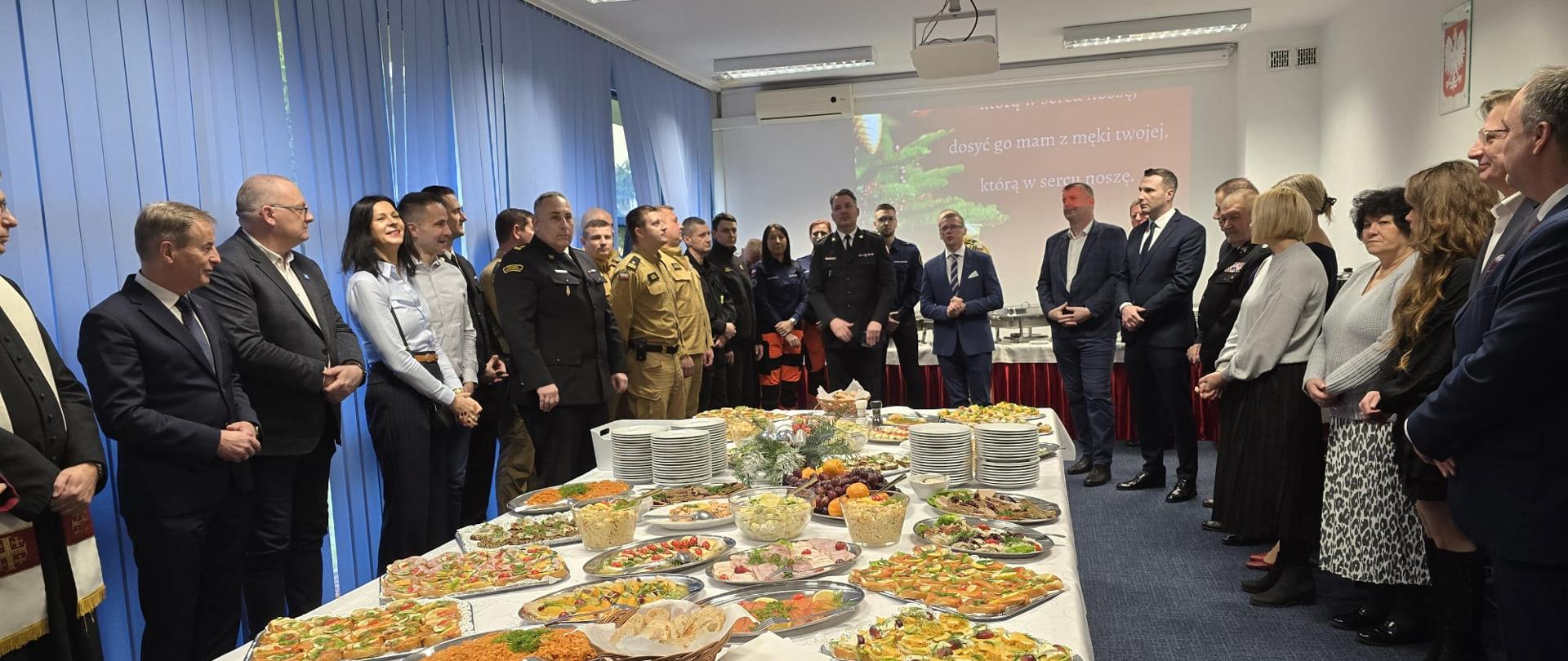 Zdjęcie przedstawia dużą grupę ludzi zgromadzonych w sali konferencyjnej na uroczystości, która jest spotkaniem opłatkowym.
Stół Cateringowy
• W centrum kadru dominuje długi stół z cateringiem. Stół jest przykryty białym obrusem, a jego boki zasłania ciemnoczerwona (bordowa) tkanina.
• Na stole znajduje się bardzo bogaty wybór potraw, ułożonych na prostokątnych tacach, w tym:
Różnorodne kanapki i przekąski (tartinki, tortille).
Sałatki i dania warzywne (widoczne marchewki z groszkiem, sałatki jarzynowe).
Koszyk z pieczywem.
Ludzie i Ubiór
• Uczestnicy są ustawieni wzdłuż obu długich boków stołu, co nadaje scenie uroczysty i formalny charakter.
• Wśród gości widać mieszankę cywilnych strojów wizytowych (garnitury, marynarki, eleganckie sukienki/spódnice) i mundurów.
o W środkowej części grupy widocznych jest kilku mężczyzn w ciemnych i jasnych mundurach (Straży Pożarnej lub innych służb).
o Po lewej stronie, na samym skraju, widoczny jest mężczyzna w stroju duchownego (czarna sutanna i biała komża/alba na szyi),
Tło
• W pomieszczeniu panuje jasne, białe oświetlenie sufitowe.
• Po lewej stronie znajdują się długie, niebieskie zasłony.
• Na prawej ścianie wisi ekran projekcyjny, na którym widać fragment świątecznej grafiki i tekst:
"Dosyć go mam z męki twojej, która w sercu pocz..."
• Powyżej ekranu widoczne jest godło i flaga Polski na ścianie.
