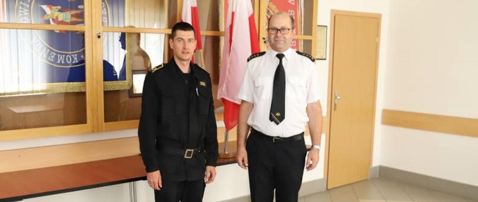 Zdjęcie przedstawia od prawej Komendanta Powiatowego Państwowej Straży Pożarnej w Zambrowie starszego brygadiera Krzysztofa Konieckiego w ubraniu letnim, wyjściowym oraz młodszego aspiranta Tomasza Kolińskiego w ubraniu dowódczo-sztabowym. Funkcjonariusze znajdują się w centralnej części zdjęcia, frontem do obiektywu. Zdjęcie zostało zrobione z okazji mianowania na wyższe stanowisko służbowe młodszego aspiranta Tomasza Kolińskiego i wykonane w jednym z pomieszczeń Komendy Powiatowej Państwowej Straży Pożarnej w Zambrowie.