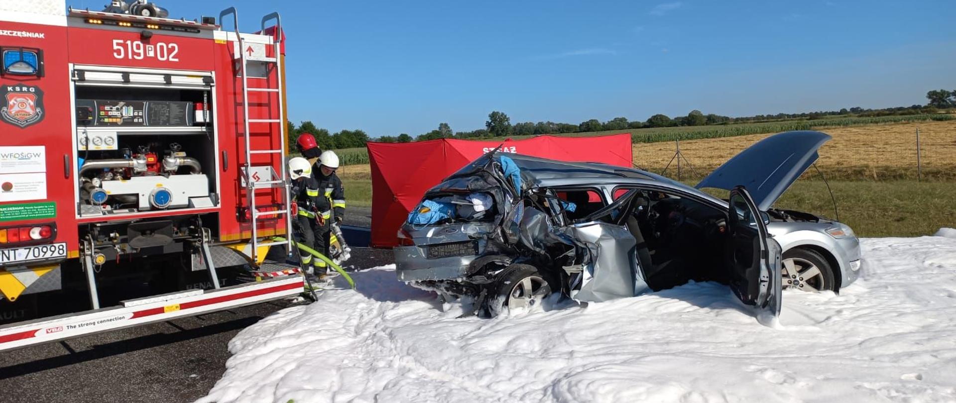 Samochód osobowy z rozbitym tyłem, duża ilość piany gaśniczej na powierzchni drogi, obok stoi samochód pożarniczy i strażacy, w tle parawan osłaniający.