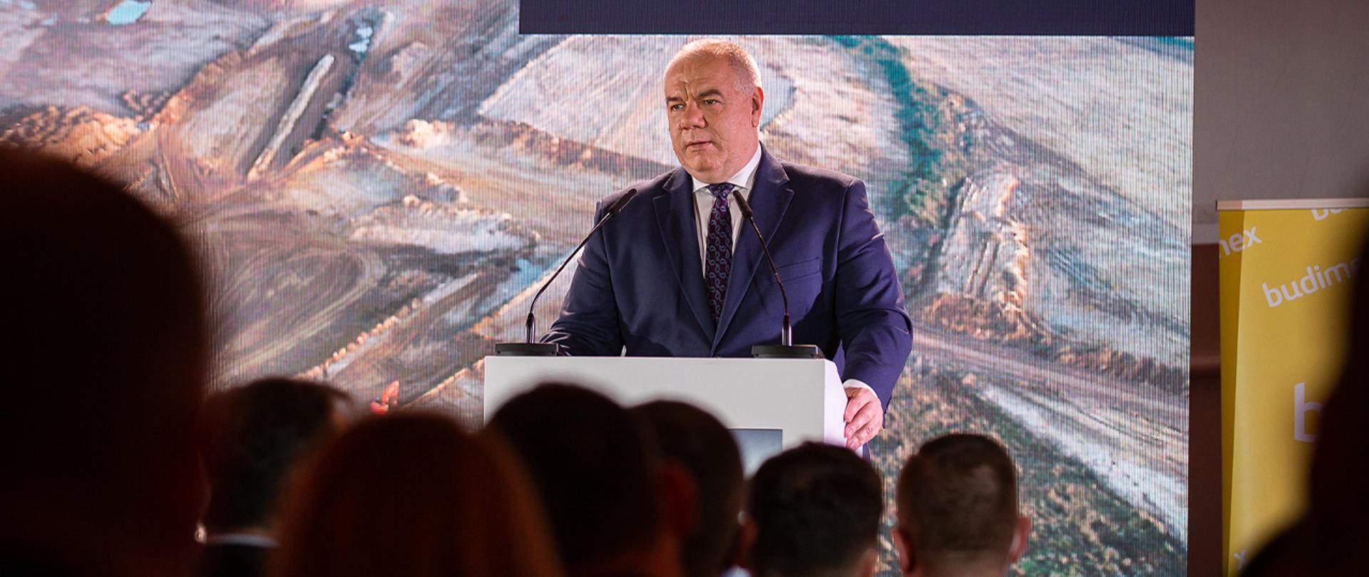 Wiceminister przy mównicy, w tle na telebimie wyświetla się zdjęcie inwestycji