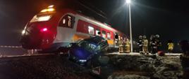 Widać pociąg osobowy, na torowisku jest samochód osobowy, w oddali po prawej stronie kilku strażaków