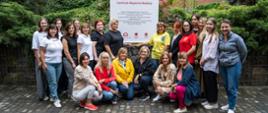 A group of 21 women posing for photograph next to a banner displaying information regarding the funding