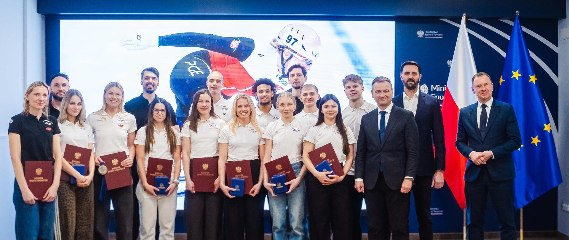 Spotkanie Sławomira Nitrasa z zawodnikami w łyżwiarstwie szybkim oraz short tracku. W tle ekran oraz flagi Polski i Unii Europejskiej