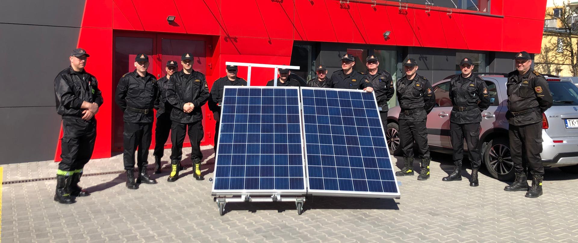 Strażacy ubrani w ubranie koszarowe stoją za trenżerem panela fotowoltaicznego na tle budynku krajowej bazy sprzętu specjalistycznego.