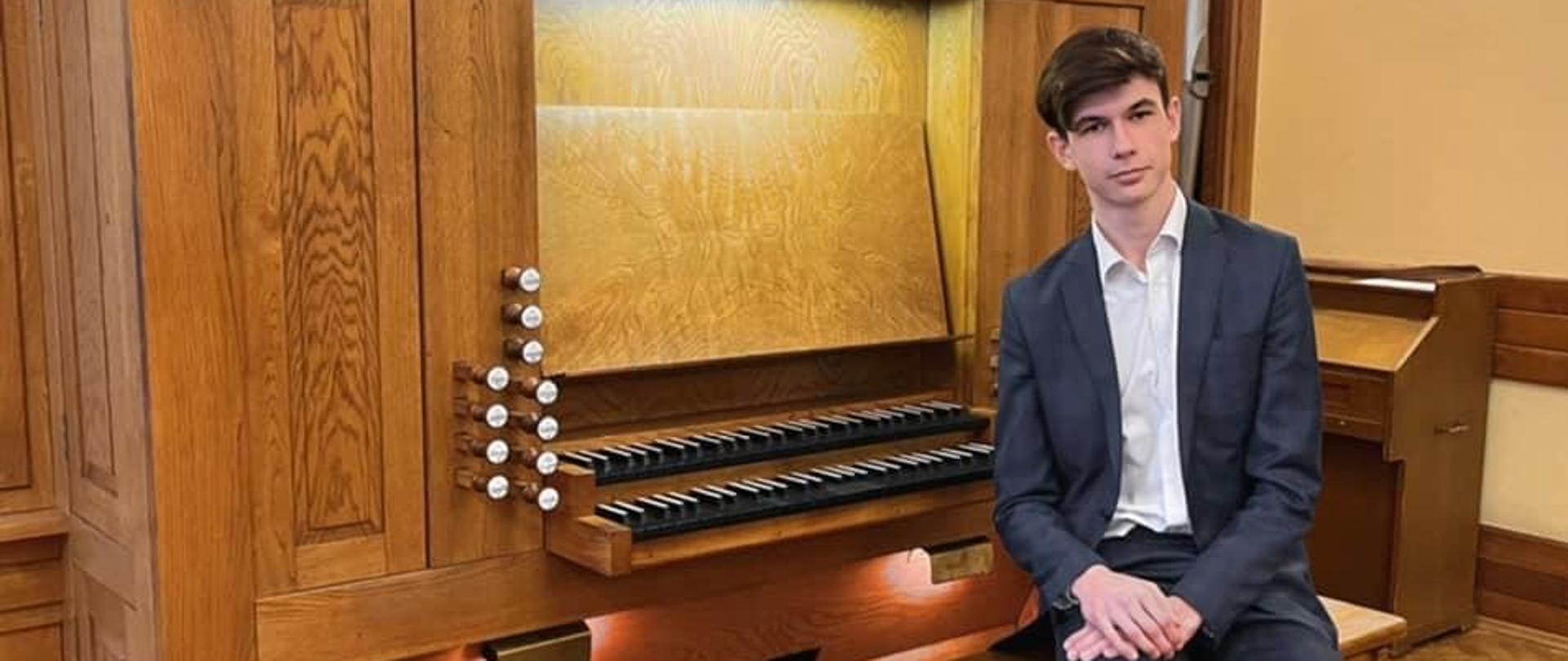 Uczeń siedzi przy organach w stroju galowym, za nim 2 manuały w kolorze czarnym