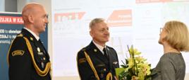 Komendanci wręczają pani kwiaty. Za nimi w tle slajd z prezentacji.