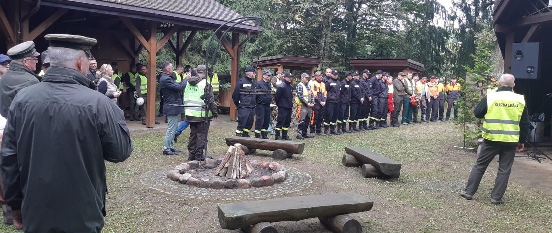 Zdjęcie przedstawia uczestników turnieju drwali podczas podsumowania zawodów na terenie polany leśnej 