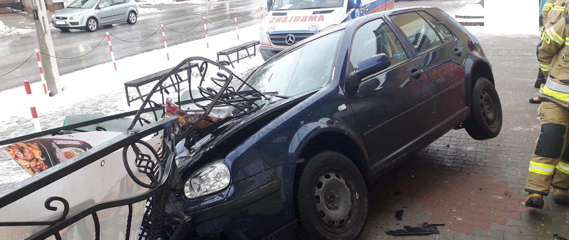 Zdjęcie przedstawia samochód osobowy koloru ciemnego, który wjechał w bariery przy sklepie spożywczym. W tle karetka pogotowia.
