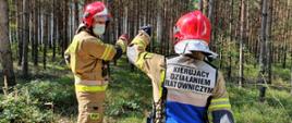 4. Zdjęcie przedstawia dwóch strażaków w ubraniach specjalnych z czerwonymi hełmami na głowach. Jeden z nich ma założona kamizelkę "Kierujący Działaniem Ratowniczym". Strażacy wskazują na zadymienie w środku lasu.