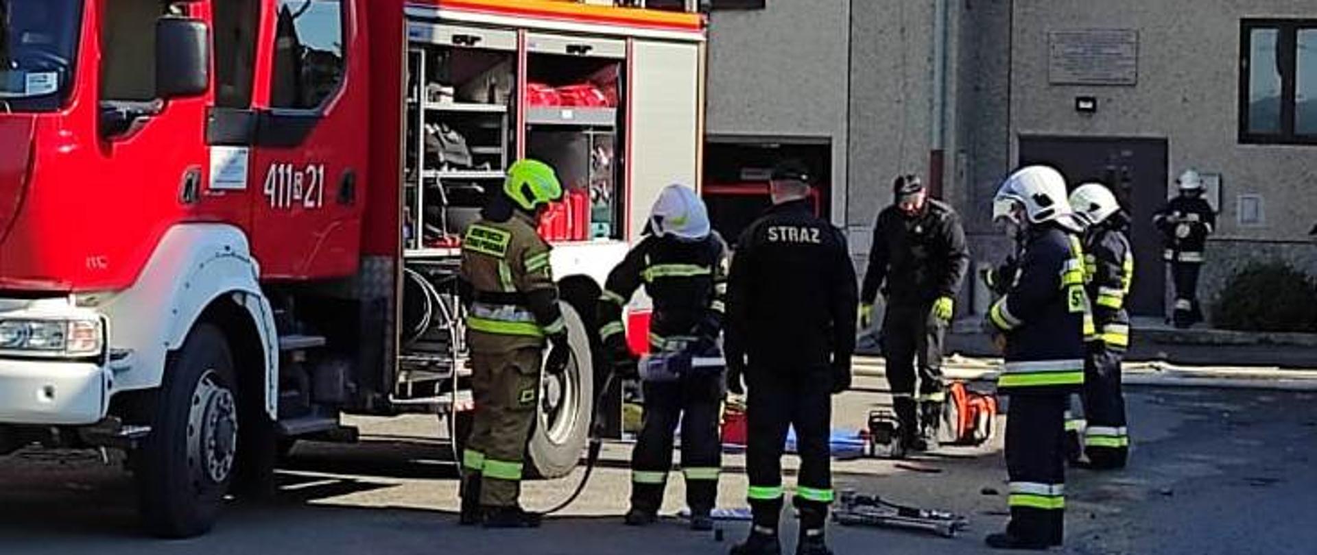 Kolorowe zdjęcie wykonane w przestrzeni otwartej w dzień. Przedstawia na pierwszym planie grupę umundurowanych na bojowo w hełmach na głowach strażaków, ćwiczących z ratowniczym sprzętem hydraulicznym i spalinowym. Po lewej stronie, na utwardzonym placu, stoi częściowo widoczny, czerwony samochód pożarniczy z otwartymi skrytkami na sprzęt, Za samochodami elewacja frontowa dużego szarego budynku, przed którym stoją też inni strażacy. W tle ściana dużego szarego budynku.