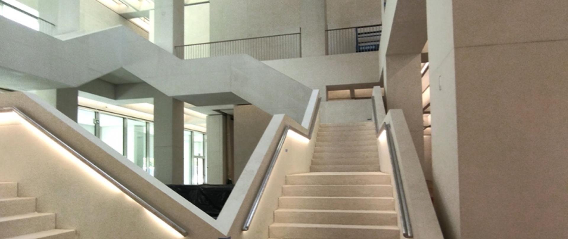 Nowoczesna klatka schodowa z jasnymi materiałami, dużymi oknami i wielopoziomową strukturą.