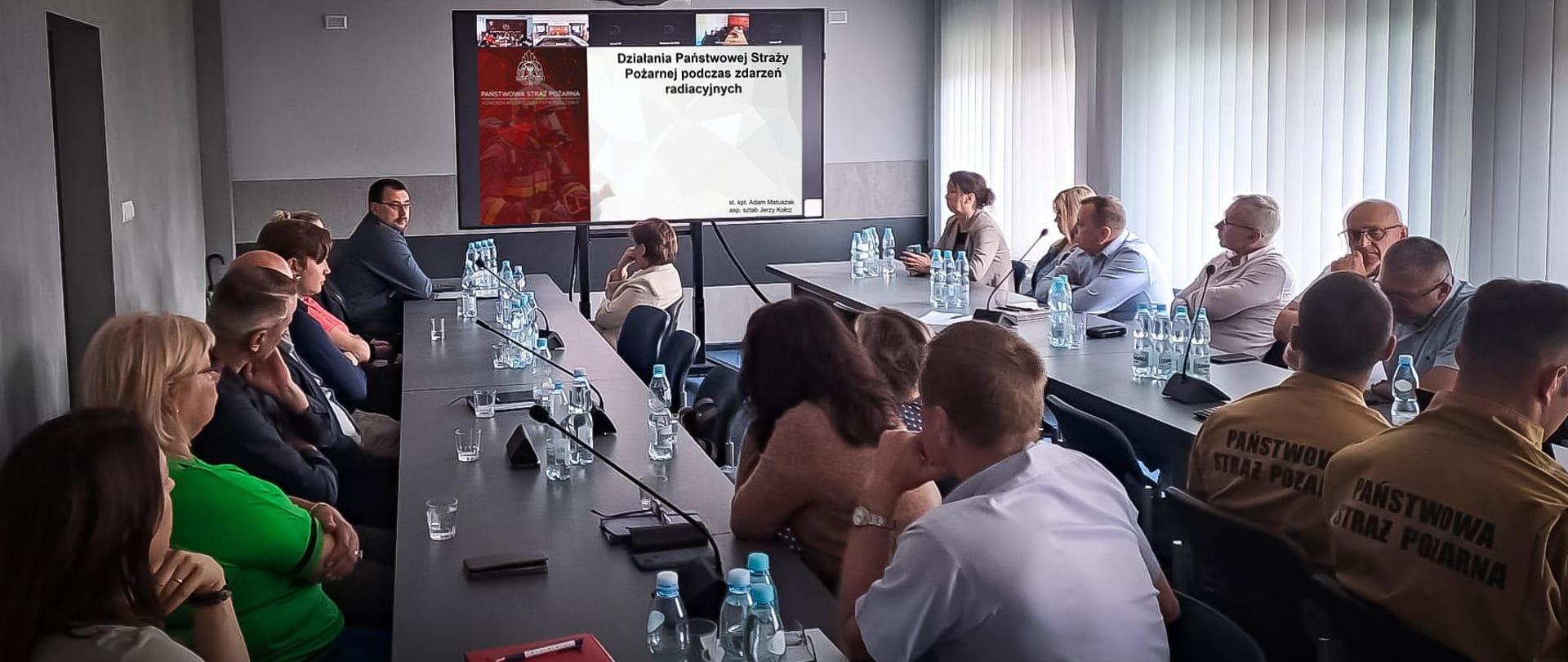 Zdjęcie zrobione w sali szkoleniowej podczas narady, w której uczestniczą osoby cywilne i funkcjonariusze PSP. Uczestnicy siedzą przy stole konferencyjnym. W tle stoi monitor z wyświetloną prezentacją.