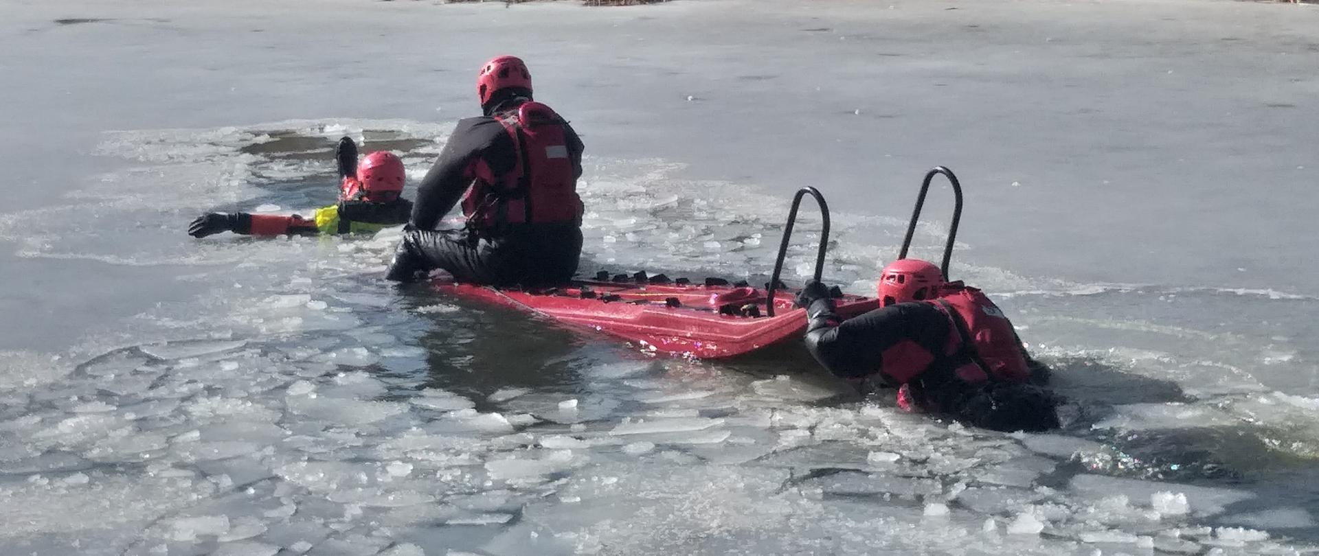 Zdjęcie przedstawia ratowników PSP zabezpieczonych w ubrania do pracy w wodzie. Strażak przy wykorzystaniu sani lodowych dociera do pozoranta oraz ewakuuje go na sanie lodowe. Akwen wodny pokryty warstwą łamiącego się lodu.