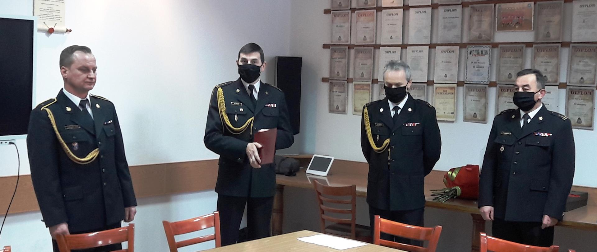 Kolorowa fotografia przedstawia zbiórkę strażaków w świetlicy z okazji przejścia na emeryturę asp. Jacka Kuśnierczyka. Na pierwszym planie stoją czterej strażacy w umundurowaniu galowym ze sznurem, bez nakrycia głowy, w czarnych maseczkach. Od lewej (bez maseczki) aspirant Jacek Kuśnierczyk, starszy brygadier Krzysztof Folta, młodszy brygadier Bogdan Biedka, brygadier Krzysztof Szerszeń. W tle jasne ściany świetlicy z widocznymi licznymi dyplomami.