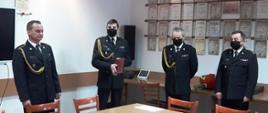 Kolorowa fotografia przedstawia zbiórkę strażaków w świetlicy z okazji przejścia na emeryturę asp. Jacka Kuśnierczyka. Na pierwszym planie stoją czterej strażacy w umundurowaniu galowym ze sznurem, bez nakrycia głowy, w czarnych maseczkach. Od lewej (bez maseczki) aspirant Jacek Kuśnierczyk, starszy brygadier Krzysztof Folta, młodszy brygadier Bogdan Biedka, brygadier Krzysztof Szerszeń. W tle jasne ściany świetlicy z widocznymi licznymi dyplomami.
