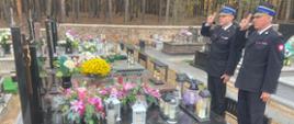 Na zdjęciu widać strażaków oddających hołd zmarłemu byłemu funkcjonariuszowi straży pożarnej. W tle widać inne pomniki znajdujące się na cmentarzu oraz roślinność.