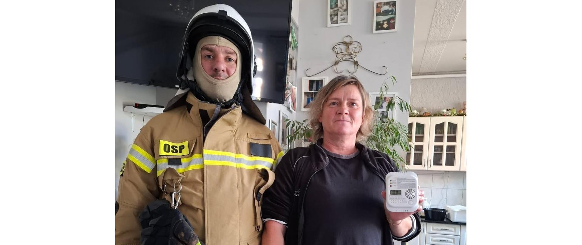 Zdjęcie przedstawia strażaka Ochotniczej Straży Pożarnej w Grabowie, stojący obok mieszkanki lokalu, w którym uruchomiła się czujka tlenku węgla alarmując o zagrożeniu. Kobieta trzyma w lewej dłoni czujkę tlenku węgla