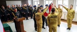 Uroczyste ślubowanie strażaków. Czterech funkcjonariuszy w piaskowych mundurach stoi przy sztandarze, podnosząc dwa palce prawej dłoni w geście przysięgi. Przy mównicy stoi komendant, a w tle zgromadzeni oficerowie obserwują ceremonię.