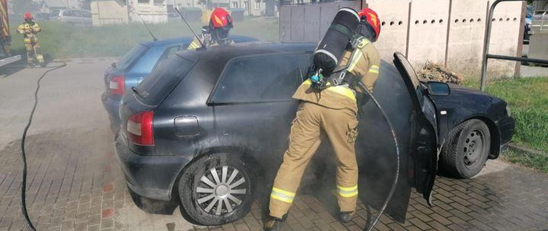 Na zdjęciu czarne auto osobowe z którego wydobywa się dym. Przy aucie strażacy w mundurach, czerwonych hełmach i aparatach powietrznych na plecach. Gaszą pożar samochodu. W tle bloki mieszkalne.