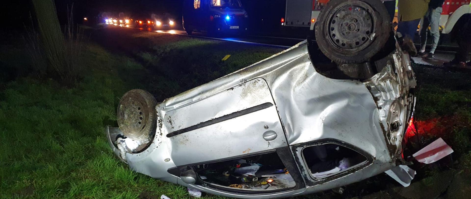 Pora nocna. Rów obok drogi. Zniszczone srebrne auto osobowe leży na dachu na zielonej trawie i brązowej ziemi. W tle czerwony wóz pożarniczy oraz biały ambulans pogotowia ratunkowego z włączonymi światłami oraz inne pojazdy osobowe.