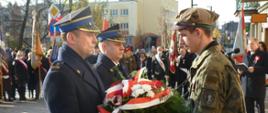 Dwóch mężczyzn w mundurach wyjściowych straży pożarnej w tym komendant powiatowy brygadier Mariusz Wojcieszko składa wiązankę z biało-czerwonymi szarfami na ręce mężczyzny ubranego w mundur wojskowy. W tle uczestnicy uroczystości.