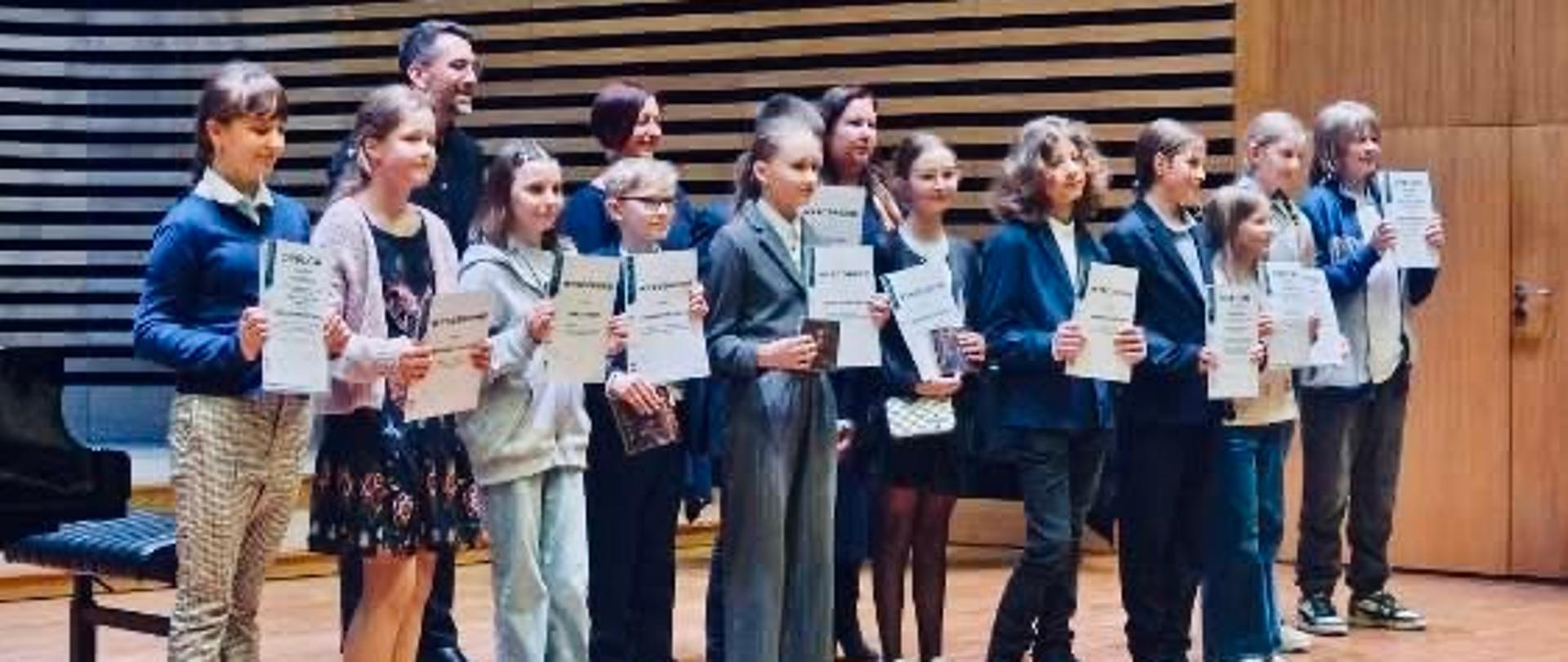 Grupa dzieci i młodzieży stojąca na scenie w sali koncertowej, trzymająca dyplomy. Uczestnicy ustawieni są w dwóch rzędach na drewnianej podłodze sceny. Ubrani są w eleganckie stroje - garnitury, sukienki i spodnie. W tle widoczna jest nowoczesna akustyczna ściana z drewnianych paneli ułożonych w poziome pasy. Po lewej stronie sceny znajduje się fortepian. Oświetlenie sceniczne podkreśla uroczysty charakter wydarzenia.
