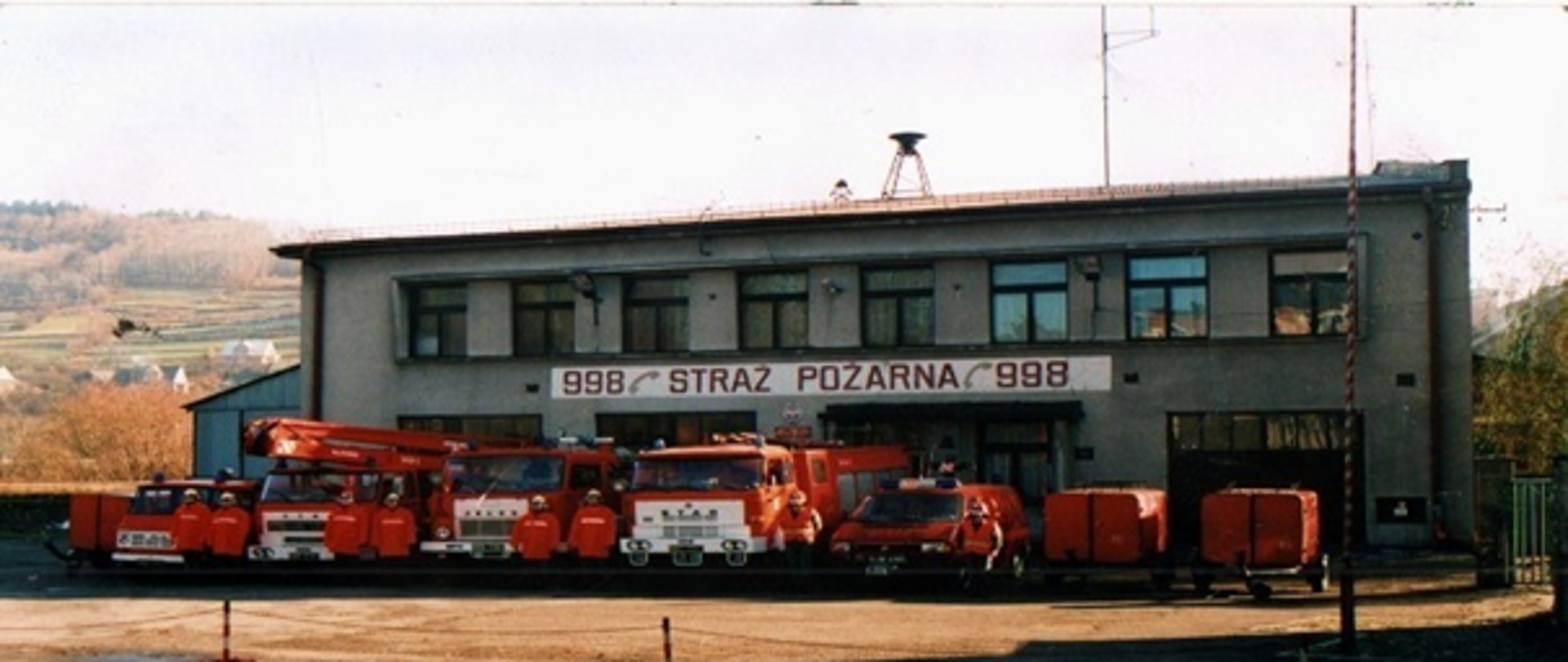 Zdjęcie przedstawia siedzibę Jednostki Ratowniczo - Gaśniczej w Suchej Beskidzkiej. Zdjęcie przedstawia budynek, przed budynkiem stoją wozy strażackie z lat 90.