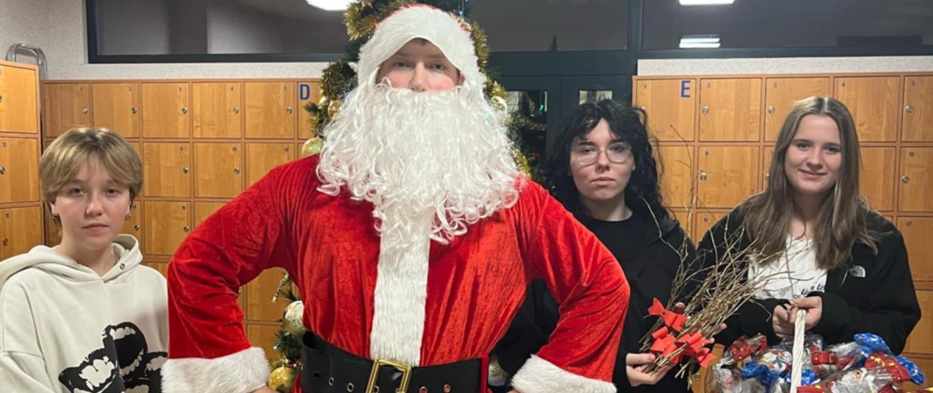 Na zdjęciu stoi chłopiec przebrany w strój świętego Mikołaja. Po lewej stronie stoi dziewczyna trzymająca granatową teczkę z kartkami, po prawej stoją dwie dziewczyny, jedna trzyma rózgi, a druga biały koszyk ze słodyczami. W tle widać fragmenty choinki i szafek.