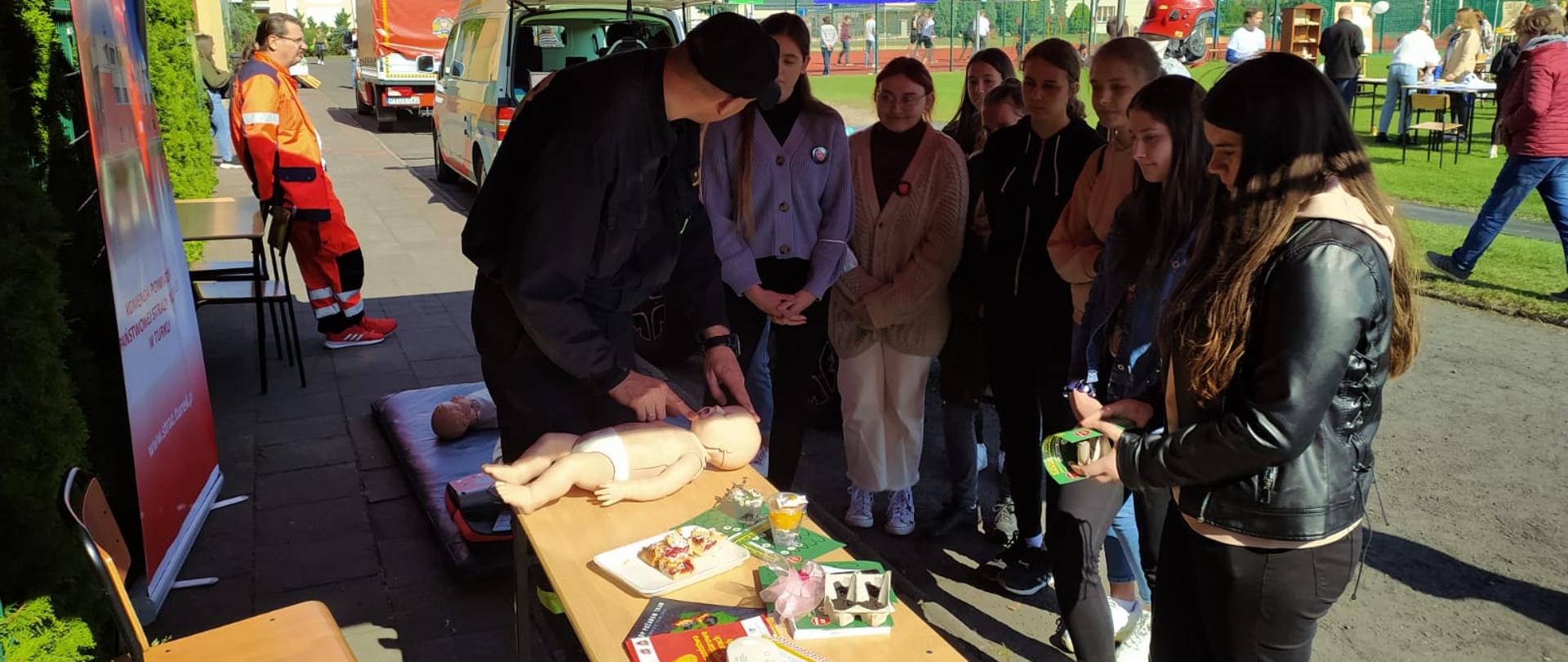 Strażak przedstawia jak prawidłowo prowadzić resuscytacje u dziecka na manekinie. W około niego zgromadzeni słuchacze.