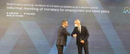 Wiceminister MRPiT Marek Niedużak stojący w masce i witający się "łokciem" z europejskim politykiem na tle baneru z angielskim napisem informującym o nieformalnym spotkaniu ministrów pracy i polityki społecznej Unii Europejskiej, które odbywa się w Brdo, w Słowenii 8-9 lipca