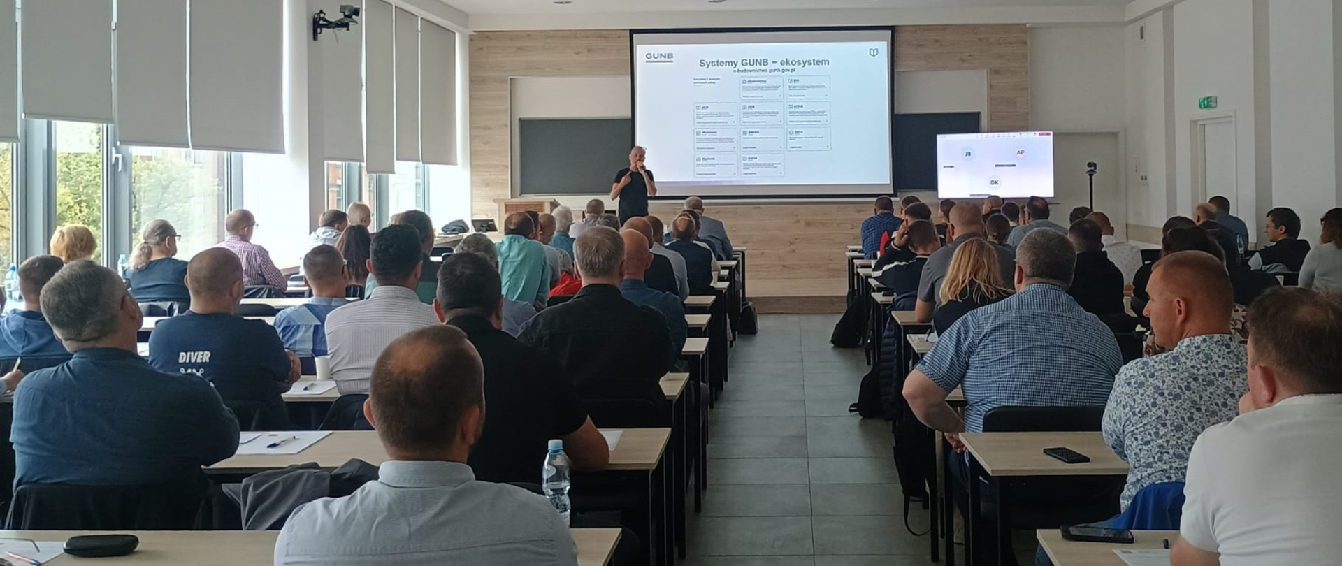 Sala szkoleniowa z dużą grupą siedzących uczestników, słuchających prowadzącego stojącego w tle przed ekranem z wyświetloną prezentacją.