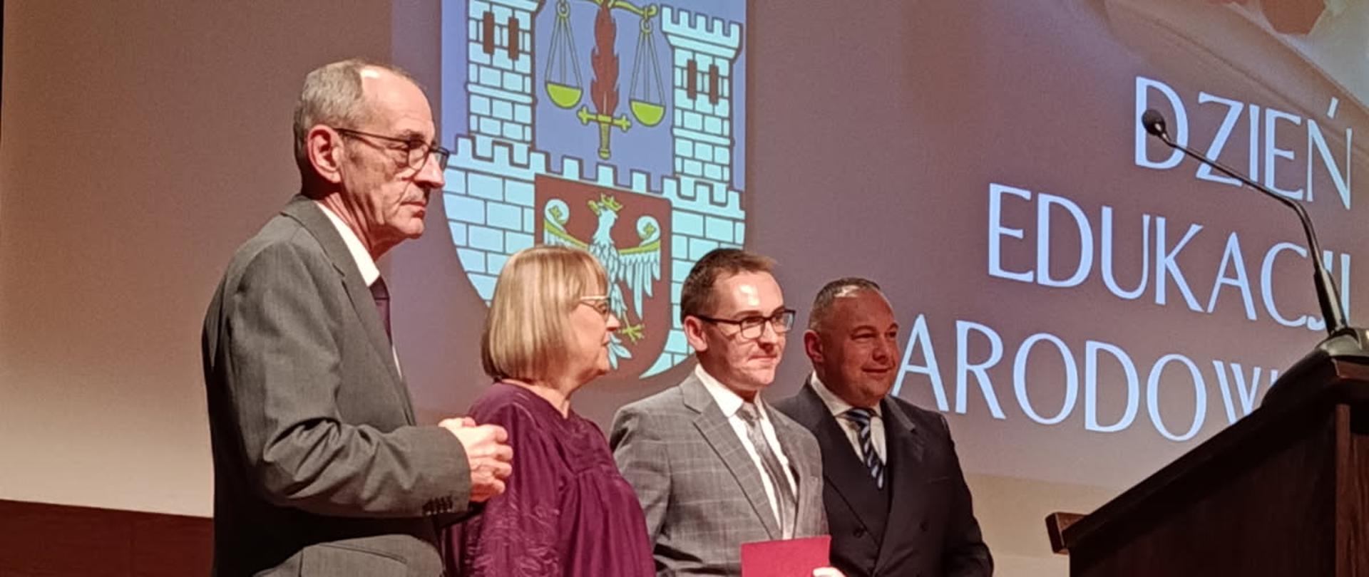  Zapytanie zostało przetworzone
Czworo dorosłych osób stoi na scenie w oświetlonej sali. Scena ma drewnianą podłogę. Po lewej stronie ekranu za nimi znajduje się herb miasta wyświetlany na dużym ekranie. Po prawej stronie ekranu wyświetlany jest tekst „DZIEŃ EDUKACJI NARODOWEJ” i otwarta książka. Po prawej stronie stoi mównica. Z lewej strony stoi mężczyzna w szarym garniturze, który trzyma rękę na ramieniu stojącej obok kobiety. Kobieta ma na sobie fioletową bluzkę i bordowe spodnie. Po prawej stronie kobiety stoi mężczyzna w szarym, wzorzystym garniturze, który trzyma w dłoni czerwony dokument, prawdopodobnie nagrodę. Obok niego, najbardziej po prawej, stoi mężczyzna w ciemnym garniturze i krawacie. W tle widoczny jest fragment fortepianu