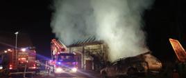 Na zdjęciu widzimy akcję ratowniczo-gaśniczą podczas pożaru. Widoczne dwa pojazdy ratownicze podczas działań. Po prawej stronie widoczny spalony samochód osobowy. Zdjęcie wykonane w porze nocnej na miejscu akcji.