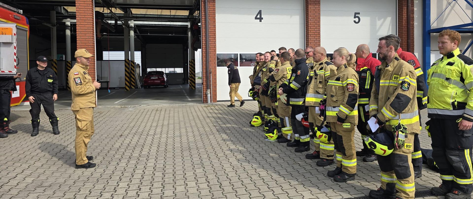 Na zdjęciu widać grupę strażaków stojących w szeregu przed budynkiem strażnicy pożarnej. Budynek ma kilka zamkniętych boksów garażowych oznaczonych numerami (widać numery 4 i 5), a w jednym z otwartych garaży stoi czerwony samochód.
Strażacy ubrani są w różne umundurowania bojowe – większość w beżowe lub czarne z odblaskowymi pasami, część w kurtkach w kolorze jaskrawożółtym. Wiele osób trzyma swoje hełmy w rękach lub pod pachą.
Po lewej stronie stoi mężczyzna w mundurze koloru piaskowego, który prowadzi odprawę do zgromadzonych strażaków. Za nim znajduje się fragment czerwonego wozu strażackiego.
Jest to oficjalna zbiórka egzaminem.
