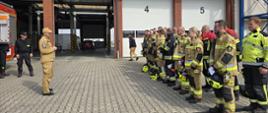 Na zdjęciu widać grupę strażaków stojących w szeregu przed budynkiem strażnicy pożarnej. Budynek ma kilka zamkniętych boksów garażowych oznaczonych numerami (widać numery 4 i 5), a w jednym z otwartych garaży stoi czerwony samochód.
Strażacy ubrani są w różne umundurowania bojowe – większość w beżowe lub czarne z odblaskowymi pasami, część w kurtkach w kolorze jaskrawożółtym. Wiele osób trzyma swoje hełmy w rękach lub pod pachą.
Po lewej stronie stoi mężczyzna w mundurze koloru piaskowego, który prowadzi odprawę do zgromadzonych strażaków. Za nim znajduje się fragment czerwonego wozu strażackiego.
Jest to oficjalna zbiórka egzaminem.
