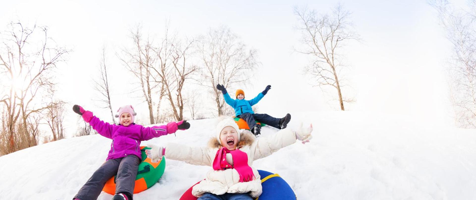 Podekscytowana grupa dzieci zjeżdżających razem na górce podczas pięknego zimowego dnia z drzewami w tle.